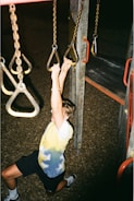 A young person hangs from playground rings, using both hands for support. They are wearing a tie-dye shirt and shorts. The playground equipment is composed of metal chains and wooden structures with red accents. The surface below is covered with wood chips.