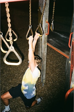A young person hangs from playground rings, using both hands for support. They are wearing a tie-dye shirt and shorts. The playground equipment is composed of metal chains and wooden structures with red accents. The surface below is covered with wood chips.