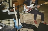 A musician is playing a white electric bass guitar while seated on a makeshift bench made of pallets. Nearby, there is a blue electric guitar resting against an amplifier. A microphone is set up in front of the musician, and various equipment, including cables and speakers, is scattered around. A bicycle wheel is partially visible in the background.