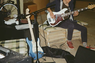 A musician is playing a white electric bass guitar while seated on a makeshift bench made of pallets. Nearby, there is a blue electric guitar resting against an amplifier. A microphone is set up in front of the musician, and various equipment, including cables and speakers, is scattered around. A bicycle wheel is partially visible in the background.