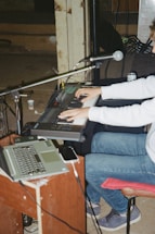 A person wearing jeans and a white sweatshirt is seated and playing an electronic keyboard. There is a microphone positioned in front, along with a laptop on a wooden stand. The setting appears to be a dimly lit room with industrial elements like a large pipe and concrete walls.