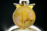 A close-up view of a gold coin with the Bitcoin symbol prominently displayed, featuring intricate circuit-like patterns on its surface. The coin appears highly detailed and is accentuated by a blurred background.