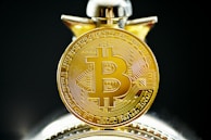 A close-up view of a gold coin with the Bitcoin symbol prominently displayed, featuring intricate circuit-like patterns on its surface. The coin appears highly detailed and is accentuated by a blurred background.