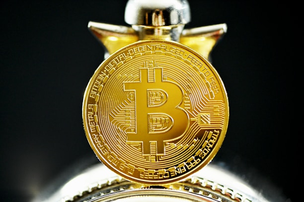 A close-up view of a gold coin with the Bitcoin symbol prominently displayed, featuring intricate circuit-like patterns on its surface. The coin appears highly detailed and is accentuated by a blurred background.