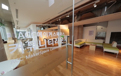 A modern, open space with wooden flooring and glass partitions, featuring a lounge area with green and beige seating. There are large windows letting in natural light, and the environment gives a clean, contemporary feel. Korean text is visible on the glass, indicating it is a MakerSpace or community lounge.