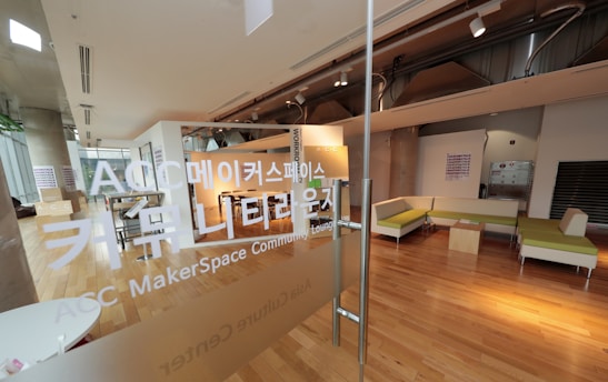 A modern, open space with wooden flooring and glass partitions, featuring a lounge area with green and beige seating. There are large windows letting in natural light, and the environment gives a clean, contemporary feel. Korean text is visible on the glass, indicating it is a MakerSpace or community lounge.