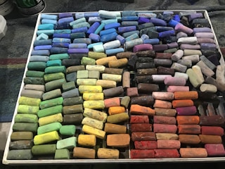 A neatly arranged collection of colorful chalk pastels is displayed in a grid-like pattern inside a white tray. Each row contains pastels of similar shades, including blues, greens, yellows, oranges, reds, pinks, purples, and browns.