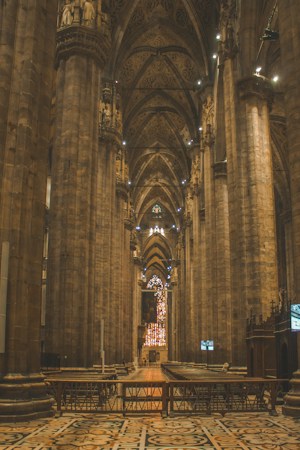 A grand interior of a Gothic cathedral with towering stone pillars and intricate vaulted ceilings. The floor is elaborately tiled, leading to a distant stained glass window glowing with warm colors. Soft lighting enhances the architectural details creating a sense of reverence and awe.