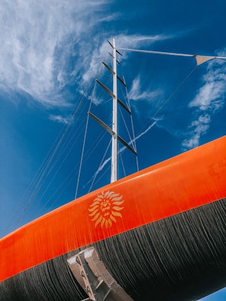 A vibrant orange sail with a lion emblem stands tall against a clear blue sky, with wispy white clouds in the background. The mast and rigging are visible, adding a nautical theme to the image.