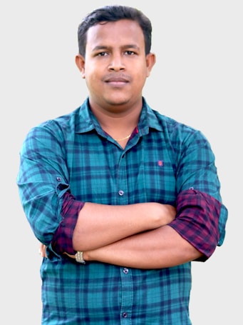 A person with short hair, wearing a green and blue checkered shirt with rolled-up sleeves, has their arms crossed and stands against a plain, light-colored background. The expression appears calm and neutral.