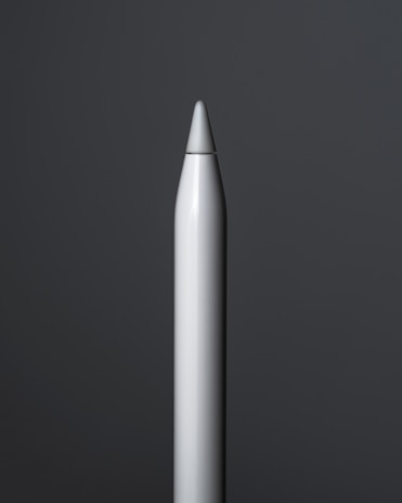 A close-up view of a sleek, metallic stylus pen standing upright against a dark grey background. The pen has a smooth, reflective surface with a pointed tip, and the lighting creates subtle highlights along its body.