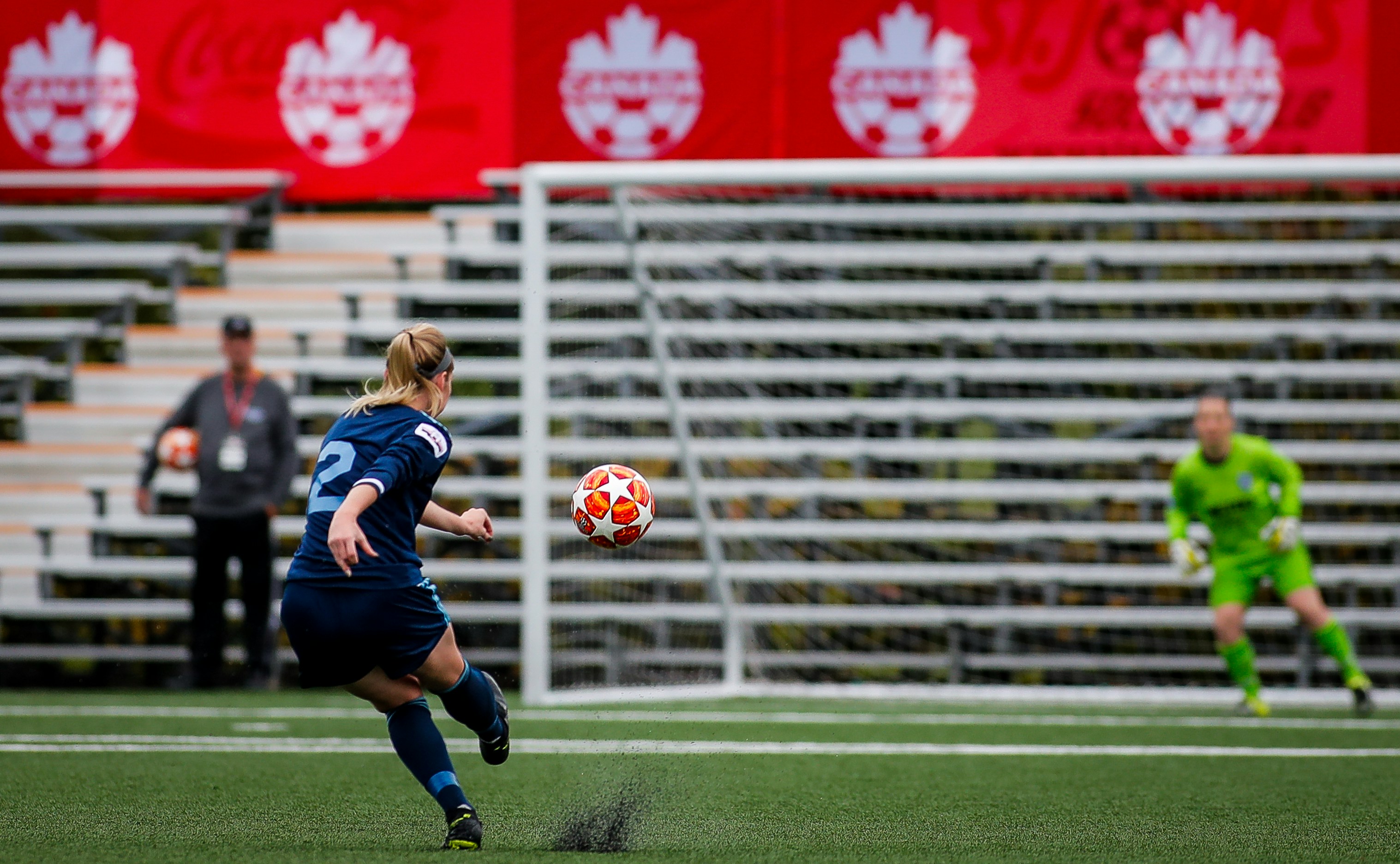 Young women shooting the soccer ball towards the goal.  The soccer goalkeeper is moving to her right in preparation to save the shot. Football Manager looks on.by My Profit Tutor