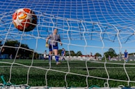 A soccer ball is captured just entering the goal from behind the net, while a goalkeeper in blue and white gear observes its path. Several soccer players wearing blue uniforms are visible on the green field, with trees and a clear blue sky in the background. The scene suggests an outdoor soccer match with a focus on a goal being made.