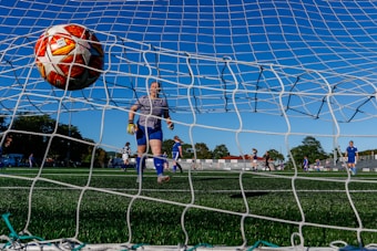 A soccer ball is captured just entering the goal from behind the net, while a goalkeeper in blue and white gear observes its path. Several soccer players wearing blue uniforms are visible on the green field, with trees and a clear blue sky in the background. The scene suggests an outdoor soccer match with a focus on a goal being made.