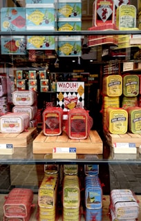A display of colorful sardine cans is arranged on shelves behind a glass window. The cans come in various vibrant colors such as red, yellow, blue, and green, and they are stacked neatly. Some cans are wrapped in red netting. A sign with bold text 'WAOUH! La Saint-Georges' is prominently featured in the center.