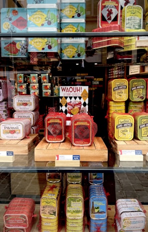 A display of colorful sardine cans is arranged on shelves behind a glass window. The cans come in various vibrant colors such as red, yellow, blue, and green, and they are stacked neatly. Some cans are wrapped in red netting. A sign with bold text 'WAOUH! La Saint-Georges' is prominently featured in the center.