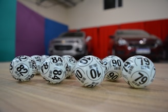 A collection of white lottery balls with black numerals scattered on a wooden surface. Two vehicles are blurred in the background, parked in a room with multicolored walls including teal, purple, blue, and red panels.