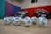 A collection of white lottery balls with black numerals scattered on a wooden surface. Two vehicles are blurred in the background, parked in a room with multicolored walls including teal, purple, blue, and red panels.