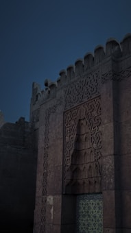 A detailed facade of a historical stone structure with intricate geometric patterns and ornate carvings. The design showcases a blend of architectural styles, with a panel featuring a repeating motif and a section of ornate blue tiles.