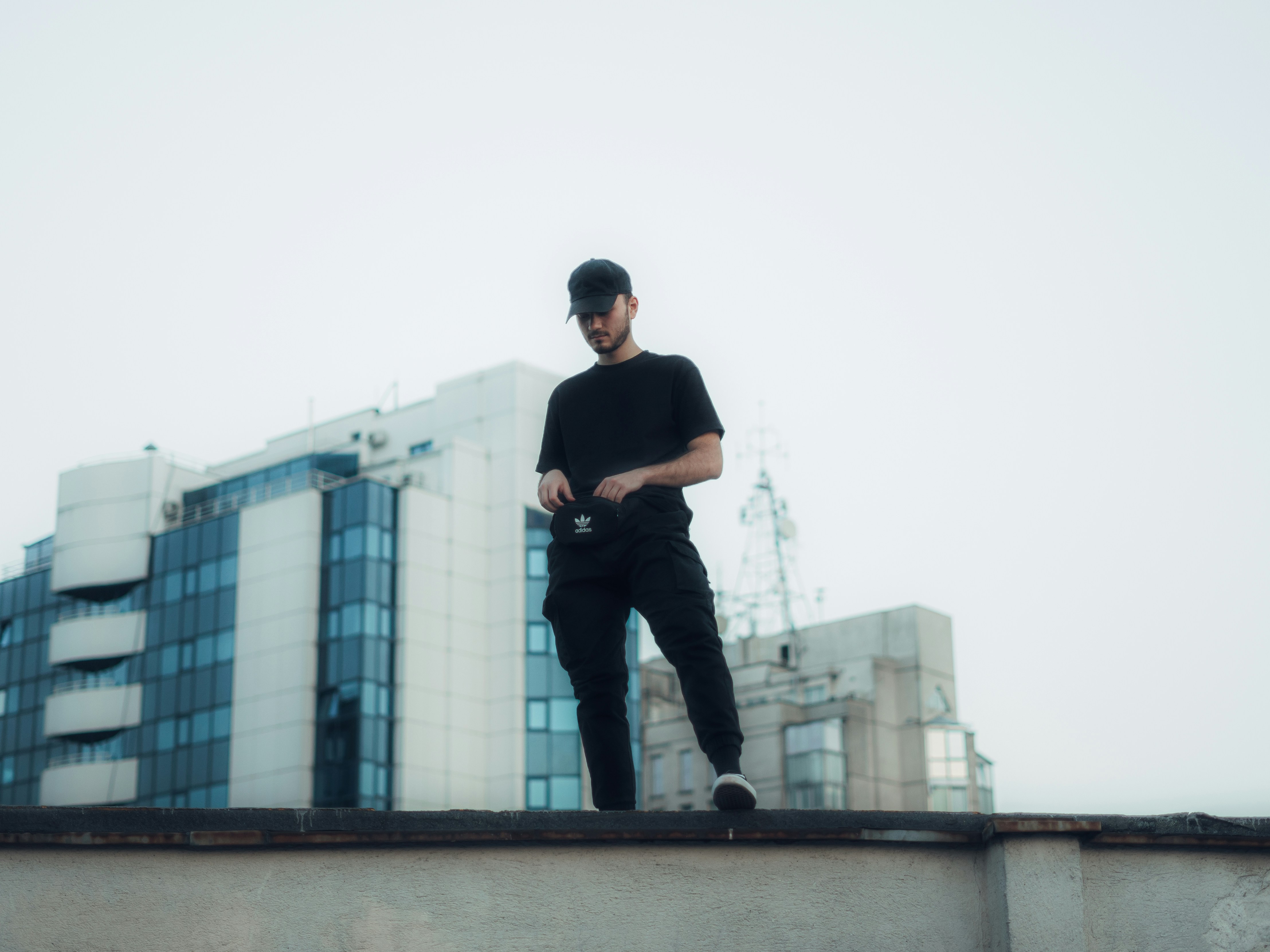 homem em jaqueta preta e jeans jeans azuis de pé na parede de concreto marrom durante o dia