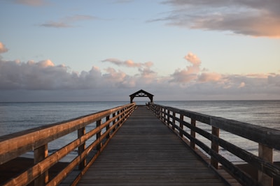 A weathered wooden pier stretching into a calm sea at dawn with pastel skies.