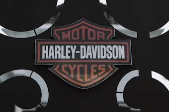 A Harley-Davidson logo is prominently displayed against a dark background. The logo features a shield shape with the words 'Harley-Davidson Motor Cycles' written in bold, with orange and white colors. Metal accents surround the logo, giving a rugged and industrial look.