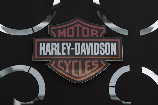 A Harley-Davidson logo is prominently displayed against a dark background. The logo features a shield shape with the words 'Harley-Davidson Motor Cycles' written in bold, with orange and white colors. Metal accents surround the logo, giving a rugged and industrial look.