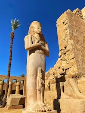 A large ancient Egyptian statue stands prominently against a clear blue sky, with a palm tree and ruins in the background. The statue depicts a figure with a traditional Egyptian headdress and crossed arms, characteristic of pharaonic imagery. The surrounding environment includes weathered stone structures and columns with remnants of hieroglyphics.
