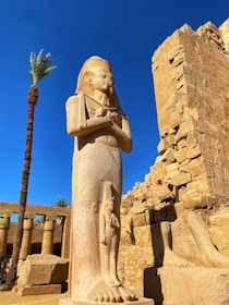 A large ancient Egyptian statue stands prominently against a clear blue sky, with a palm tree and ruins in the background. The statue depicts a figure with a traditional Egyptian headdress and crossed arms, characteristic of pharaonic imagery. The surrounding environment includes weathered stone structures and columns with remnants of hieroglyphics.