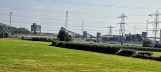 An industrial landscape with a grassy field in the foreground. Multiple transmission towers and power lines span across the image, leading to large industrial structures in the background. Trees and shrubs are scattered around the scene, adding a touch of greenery.