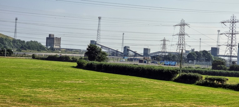 An industrial landscape with a grassy field in the foreground. Multiple transmission towers and power lines span across the image, leading to large industrial structures in the background. Trees and shrubs are scattered around the scene, adding a touch of greenery.