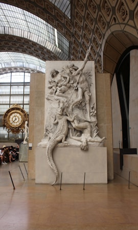 A large marble relief sculpture depicting mythological figures in a dramatic scene, including a muscular figure wielding a spear and a crocodile. The background reveals a grand architectural space with an ornate clock and arched windows.