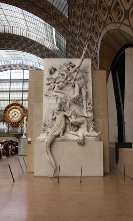 A large marble relief sculpture depicting mythological figures in a dramatic scene, including a muscular figure wielding a spear and a crocodile. The background reveals a grand architectural space with an ornate clock and arched windows.