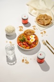 A well-arranged table setting featuring a plate of curry with boiled eggs and sliced chili peppers, accompanied by a bowl of rice and several pieces of flatbread. There are also jars with pink labels, a glass of water, and golden utensils.