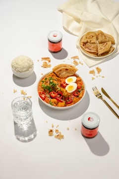 A well-arranged table setting featuring a plate of curry with boiled eggs and sliced chili peppers, accompanied by a bowl of rice and several pieces of flatbread. There are also jars with pink labels, a glass of water, and golden utensils.