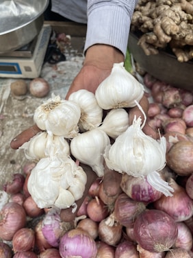 A hand holding several bulbs of garlic above a pile of red onions, with ginger in the background and a scale to the side.