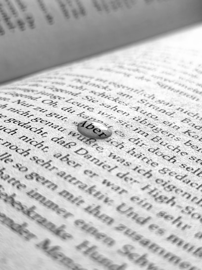 A close-up image of a book page with black printed text in German. The focus is on a small droplet or clear object resting on the page that magnifies the word 'Aber'. The background shows blurred text, suggesting a shallow depth of field.