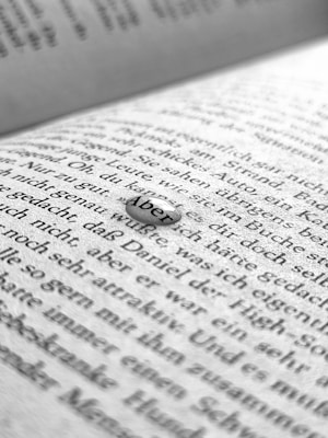 A close-up image of a book page with black printed text in German. The focus is on a small droplet or clear object resting on the page that magnifies the word 'Aber'. The background shows blurred text, suggesting a shallow depth of field.