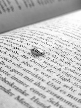 A close-up image of a book page with black printed text in German. The focus is on a small droplet or clear object resting on the page that magnifies the word 'Aber'. The background shows blurred text, suggesting a shallow depth of field.