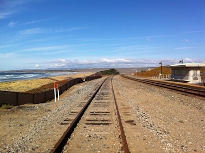 A railway track runs parallel to the coastline, with sandy and shrub-covered terrain on one side and ocean waves on the other. In the distance, a clear blue sky stretches above a horizon dotted with clouds. There is a small, modern railway structure with a flat roof nearby, along the track.