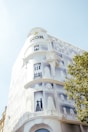 A large, ornate building with elegant architectural details features the Dior logo prominently at the top. Mannequins and silhouettes of clothing line the windows, giving an impression of a fashion display. The facade is painted with sophisticated patterns and figures. A tree is visible to the right, adding a natural element to the scene. The sky is clear and bright, suggesting a sunny day.