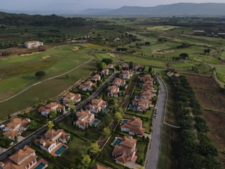 Aerial view showcasing a residential area with rows of houses featuring red-tiled roofs surrounded by green lawns and swimming pools. The community is bordered by a large, expansive golf course with neatly mowed fairways and scattered trees. In the background, lush hills and distant mountain ranges are visible under a clear blue sky.