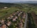 Aerial view showcasing a residential area with rows of houses featuring red-tiled roofs surrounded by green lawns and swimming pools. The community is bordered by a large, expansive golf course with neatly mowed fairways and scattered trees. In the background, lush hills and distant mountain ranges are visible under a clear blue sky.