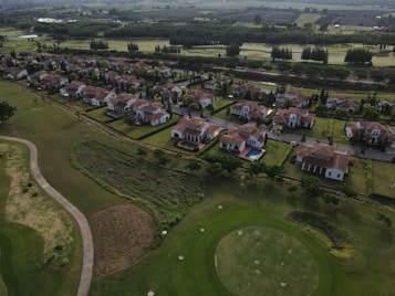 Aerial view of a suburban neighborhood featuring rows of houses with red-tiled roofs, surrounded by well-manicured lawns and trees. A golf course can be seen adjacent to the houses, with visible putting greens and sand traps. In the distance, there are lush green fields and patches of forested areas.
