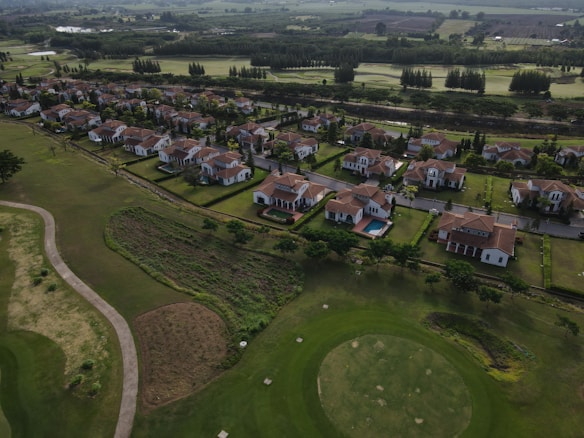 Aerial view of a suburban neighborhood featuring rows of houses with red-tiled roofs, surrounded by well-manicured lawns and trees. A golf course can be seen adjacent to the houses, with visible putting greens and sand traps. In the distance, there are lush green fields and patches of forested areas.