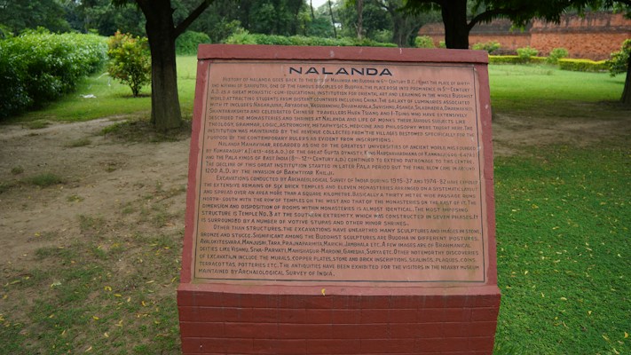 A brick plaque with detailed text about the historical site of Nalanda is situated in a lush green setting. The text provides an extensive historical background of Nalanda, an ancient center of learning. Trees and grass surround the plaque, suggesting a park or maintained garden setting.
