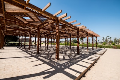 A wooden pergola structure casts intricate shadows on a paved surface. The clear blue sky and surrounding greenery create a serene atmosphere.