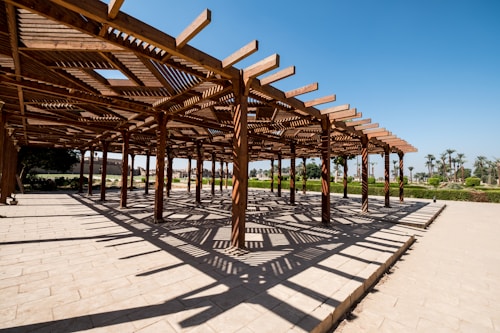A wooden pergola structure casts intricate shadows on a paved surface. The clear blue sky and surrounding greenery create a serene atmosphere.