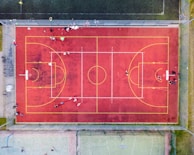An aerial view of a sports court with a vibrant red surface and yellow lines marking different sections for basketball and soccer. Several people are present on the court, some engaged in activities, while others are standing along the perimeter. The court is enclosed by a metal fence and surrounded by grass on all sides.