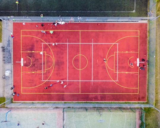 An aerial view of a sports court with a vibrant red surface and yellow lines marking different sections for basketball and soccer. Several people are present on the court, some engaged in activities, while others are standing along the perimeter. The court is enclosed by a metal fence and surrounded by grass on all sides.